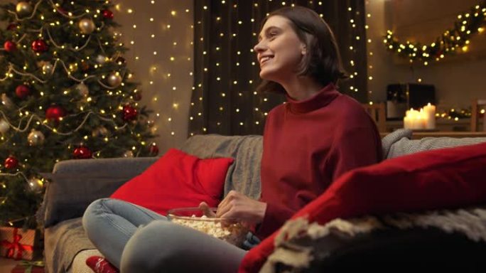 一个穿着红色毛衣的迷人的棕色头发的年轻女人正在笑着看电视，吃着爆米花。她坐在圣诞节室内的沙发上