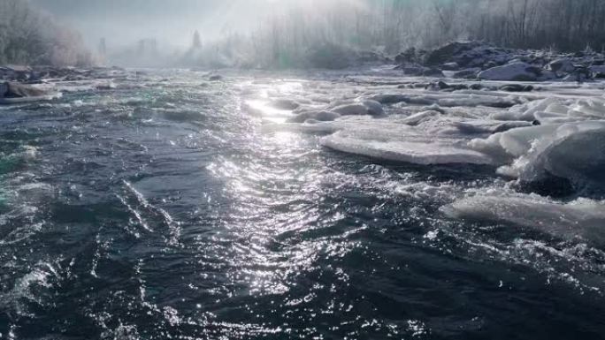 带有冰岛的山河的空中拍摄。升起的太阳的光线反射在沸腾的水流中。河流、冰和树木的冬季景观