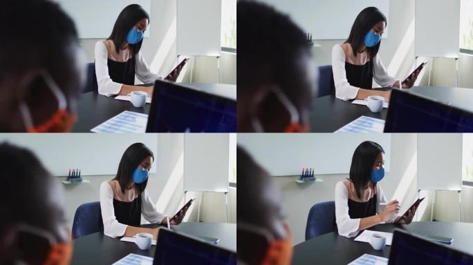 亚洲妇女在现代办公室坐在办公桌上时使用数字平板电脑戴着口罩
