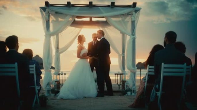 美丽的新娘和新郎在日落时在海洋海滩举行的户外婚礼上。浪漫情侣结婚、交换戒指、亲吻和与朋友分享庆祝活动