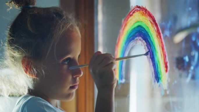 一个可爱的小创意快乐微笑的学龄前女孩画家的真实镜头正在家里厨房的窗户玻璃上画彩虹
