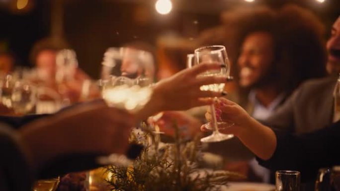 一场大型晚宴的特写镜头，一群不同的朋友在餐厅庆祝。美丽快乐的主人在晚上坐在桌子旁敬酒并举起酒杯。