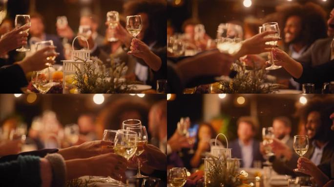 一场大型晚宴的特写镜头，一群不同的朋友在餐厅庆祝。美丽快乐的主人在晚上坐在桌子旁敬酒并举起酒杯。