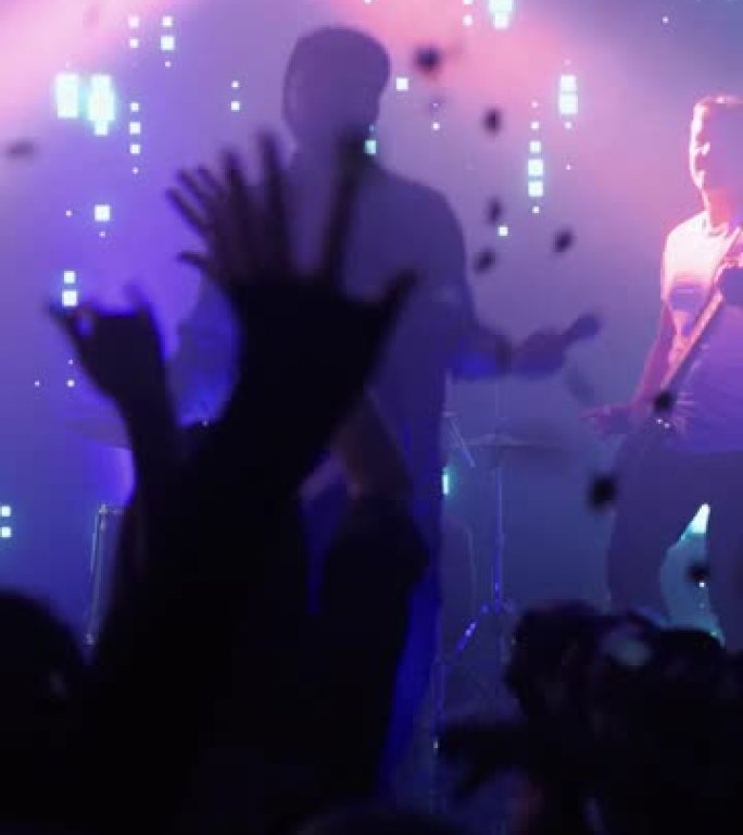 摇滚乐队在夜总会音乐会上表演的垂直镜头。前排人群正在聚会。歌迷的剪影在舞台上明亮的彩色频闪灯前举手。