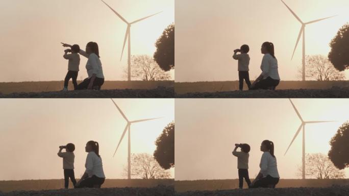家庭和母亲给2岁男婴教学的剪影日落，手持双筒望远镜看着乡村，而风力涡轮机则回到背景。教孩子们关于可持