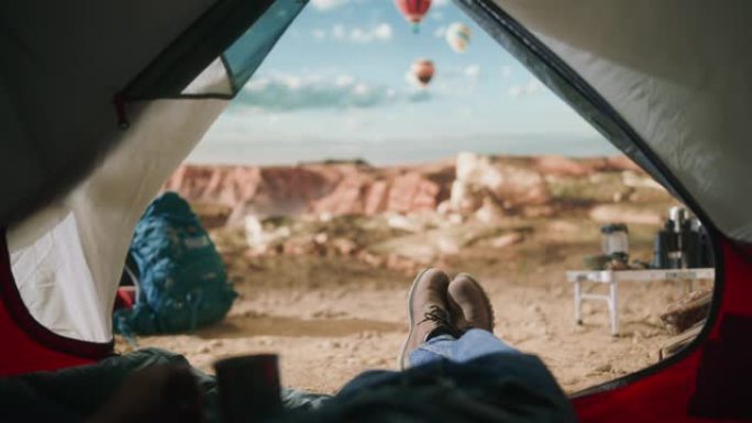 POV画面显示，一名游客在落基山顶上的户外帐篷里休息，那里有峡谷谷，并在天空中飞行热气球。背包客穿着