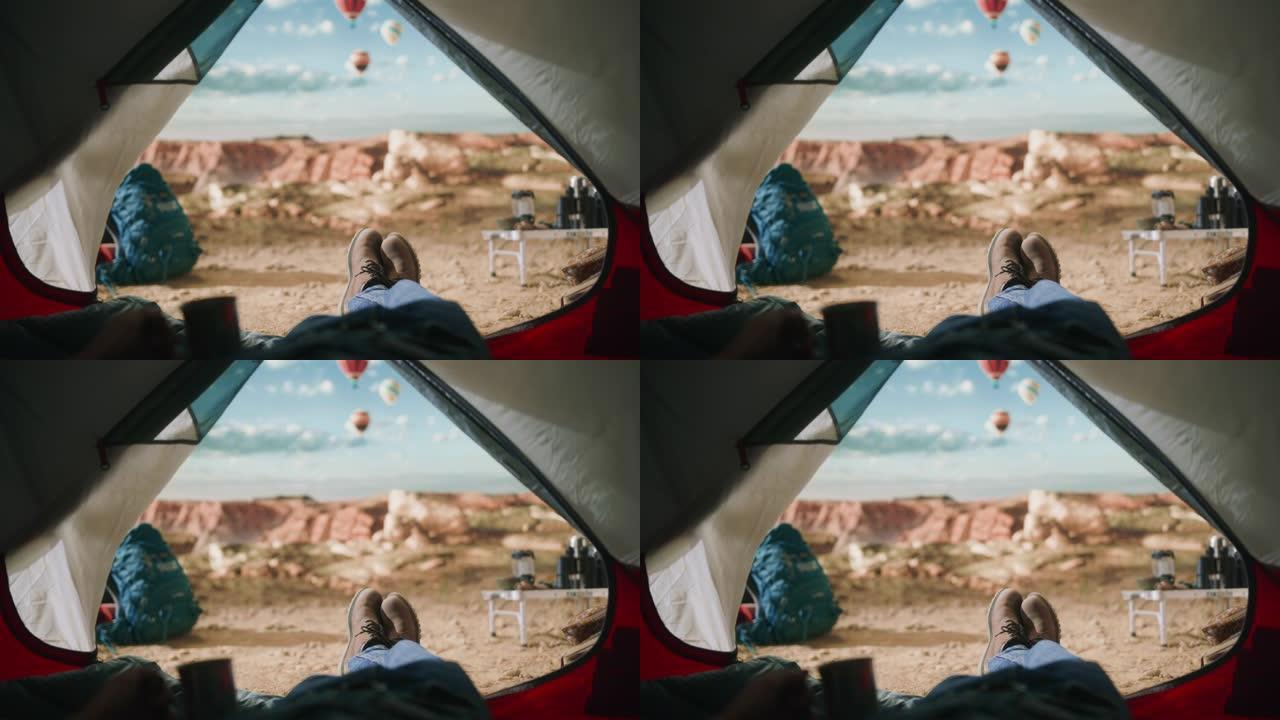 POV画面显示，一名游客在落基山顶上的户外帐篷里休息，那里有峡谷谷，并在天空中飞行热气球。背包客穿着