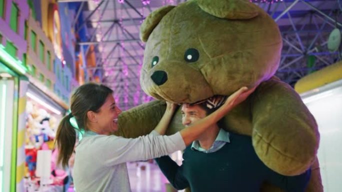 幸福无忧无虑的微笑情侣恋爱的真实镜头是与巨大的peluche泰迪熊在游乐园玩公平的射击游戏赢得了晚上