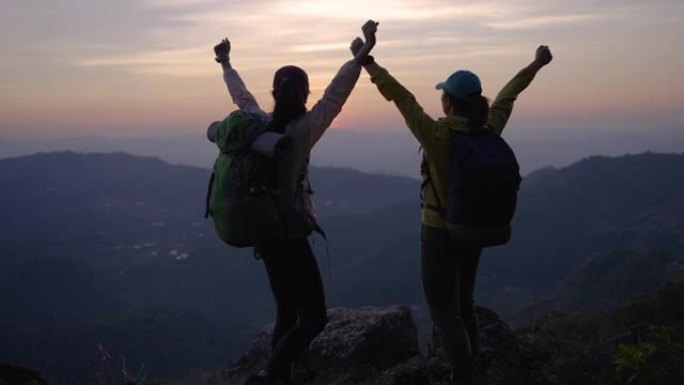 两个女人徒步上山，在山顶上击掌，举手庆祝成功