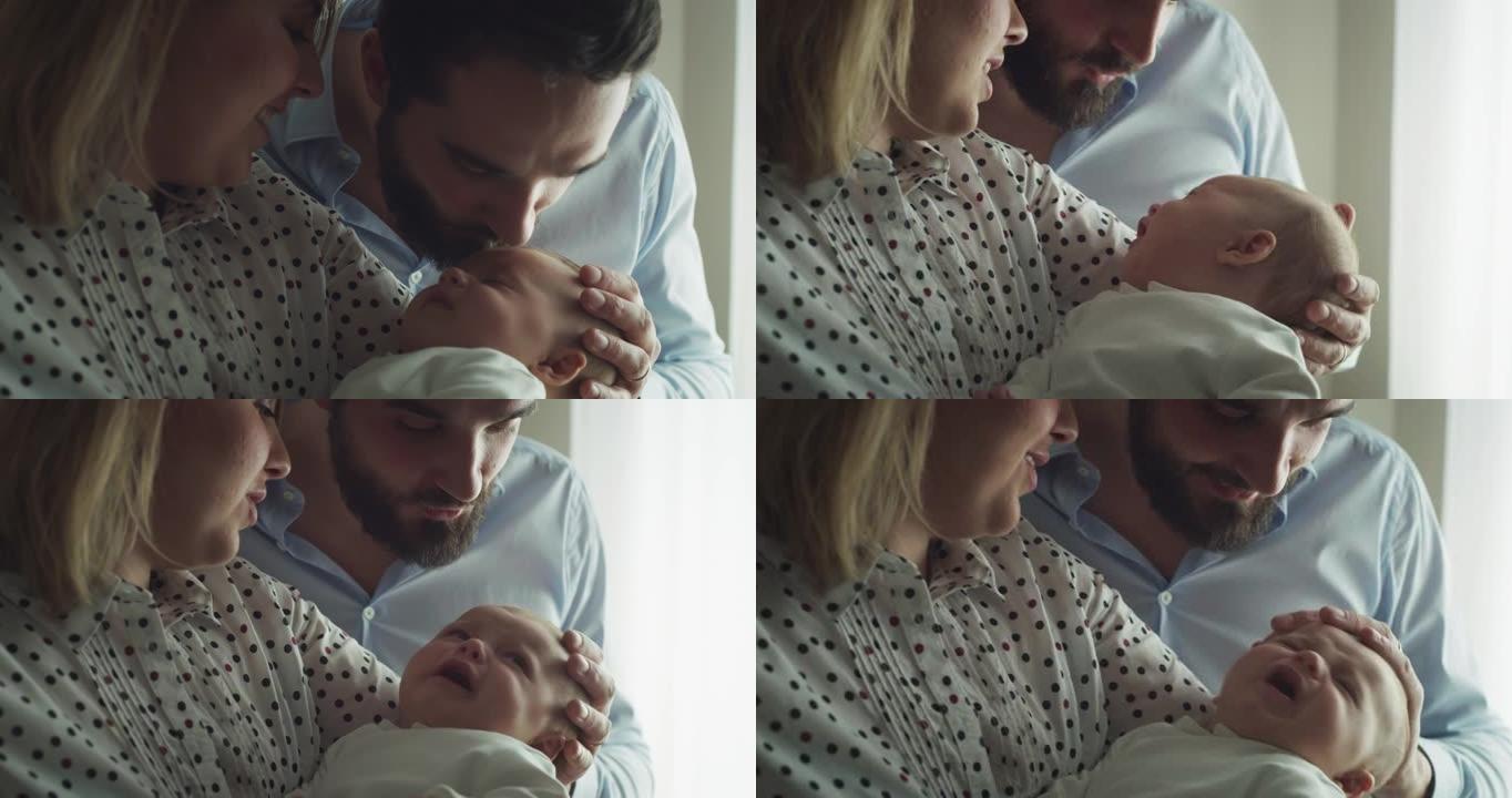 年轻父亲爱抚和亲吻他的新生婴儿的真实特写镜头，母亲早晨在家里的厨房里怀抱。家庭、父亲、母亲、父母、童