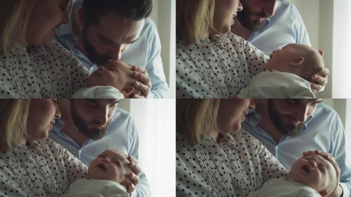 年轻父亲爱抚和亲吻他的新生婴儿的真实特写镜头，母亲早晨在家里的厨房里怀抱。家庭、父亲、母亲、父母、童