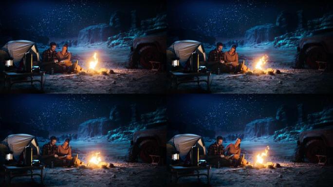 快乐情侣帐篷晚上在峡谷露营，使用坐在篝火旁的数字平板电脑。两个人发布鼓舞人心的旅行建议，在社交媒体上