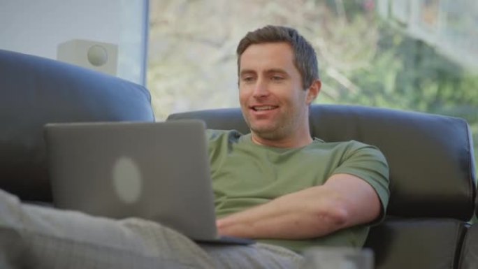 穿着睡衣的放松男子躺在家里的沙发上，用慢动作拍摄的笔记本电脑进行视频通话
