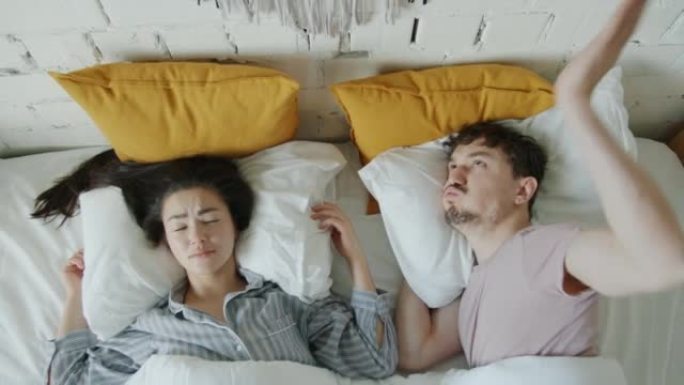 愤怒的男人和女人试图在嘈杂的公寓里睡觉，用枕头撞墙覆盖耳朵