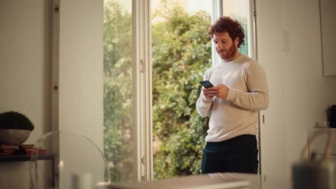 休闲成年男子，生姜卷发，使用智能手机，站在家里的客厅。英俊的男性喜欢社交媒体，在线购物，观看视频或撰