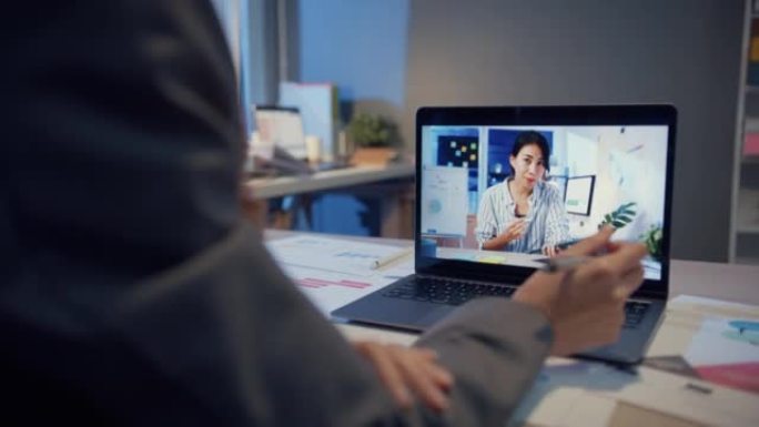 使用笔记本电脑的亚洲女性与同事谈论视频通话中的工作，而晚上在家在客厅工作。自我隔离，社会疏远，冠状病