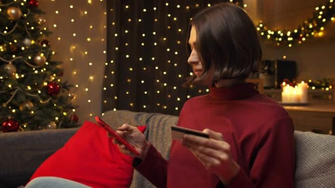一个穿着红色毛衣的可爱年轻女子正在买东西，在她的红色智能手机中输入她的卡号，她坐在圣诞节装饰的房间的