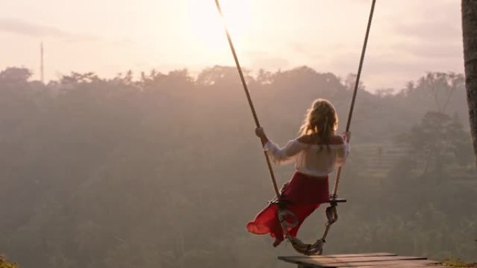 旅行女人在日出时在热带雨林上摇摆女游客坐在秋千上欣赏风景享受假期自由享受快乐假期生活方式慢动作