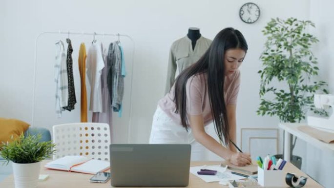 年轻的亚洲女性时装设计师使用笔记本电脑并为手工服装选择材料