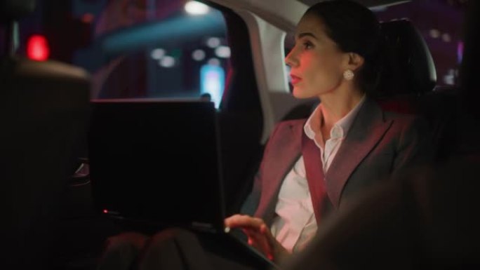 成功的女商人晚上坐在汽车后座上下班。在带有霓虹灯的城市街道上换乘出租车时，在笔记本电脑上工作的企业家