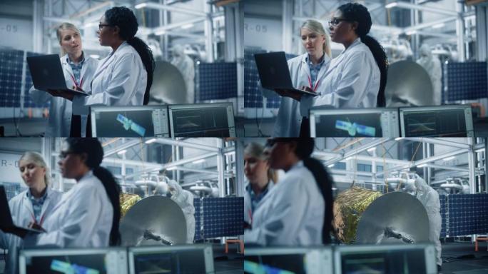 两名女工程师交谈，使用计算机在航空航天卫星制造工厂工作。由多民族科学家组成的多元化团队为太空探索计划