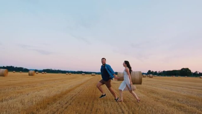 在慢动作的日落中，成年夫妇在装满吨包的田野上奔跑的跟踪镜头