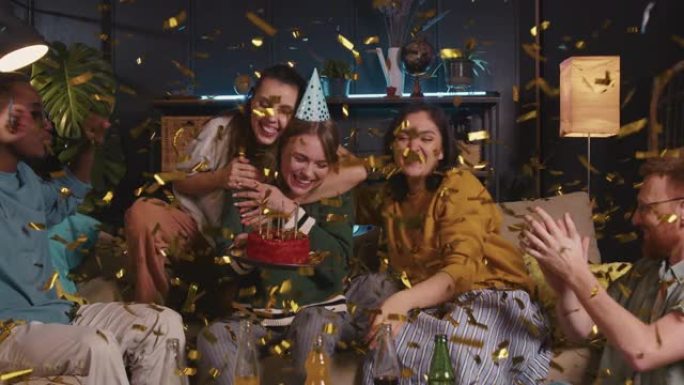 快乐兴奋美丽的金发女人在金色五彩纸屑下吹蛋糕，与有趣的不同朋友庆祝生日。