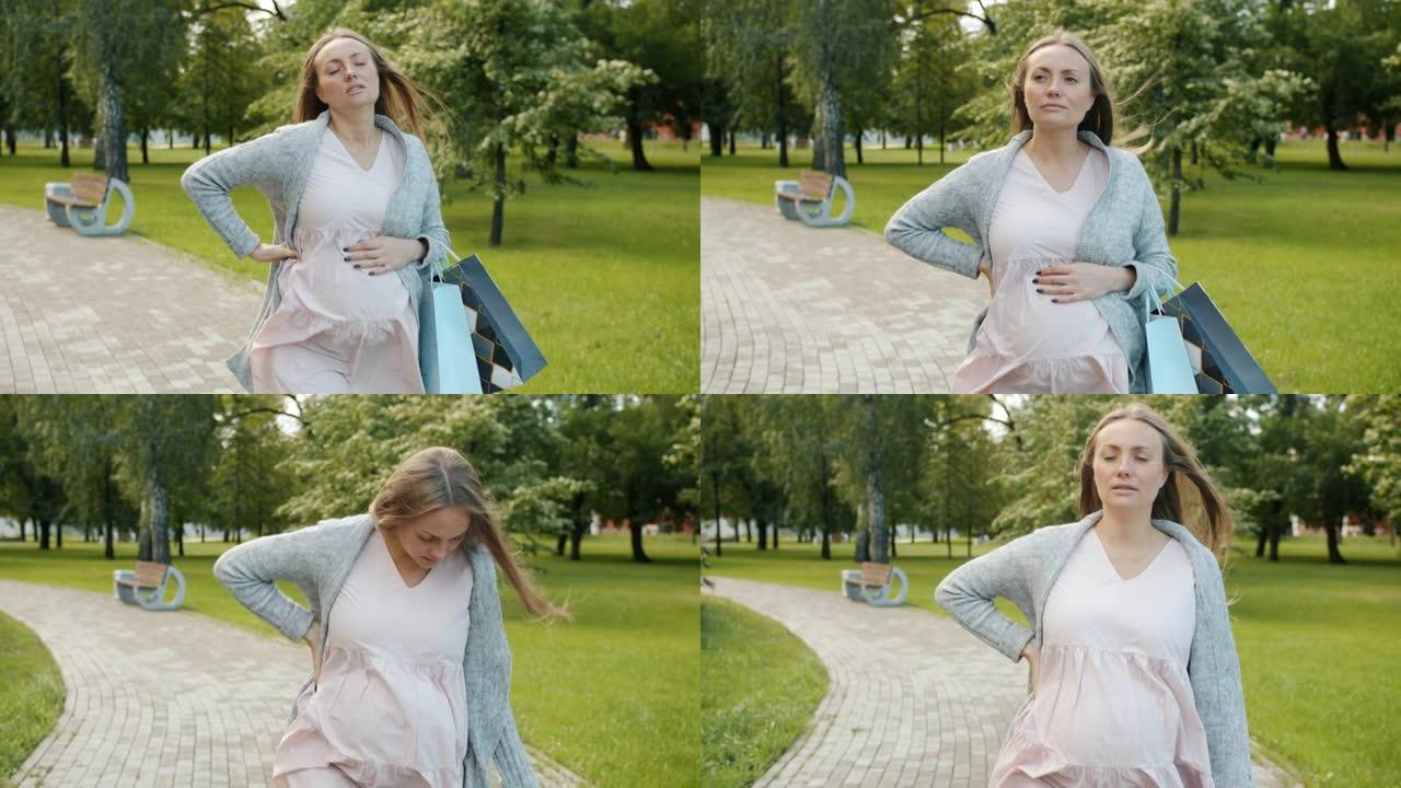 疲倦的孕妇拿着购物袋在公园散步，然后因背痛而向后抚摸