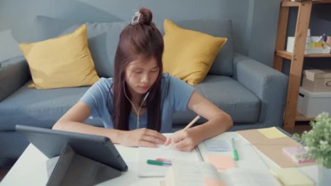 年轻的亚洲女孩少年，戴着休闲装耳机，使用数字平板电脑在线学习在家客厅写讲座笔记本。隔离教育在线电子学