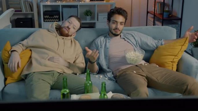 两个男人朋友晚上躺在肮脏公寓的沙发上看电视