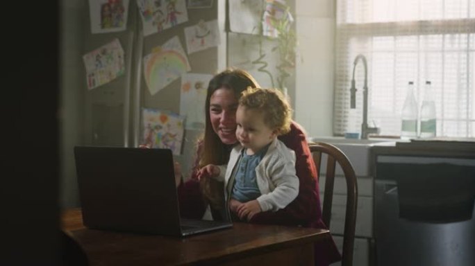 年轻快乐的母亲和她微笑的蹒跚学步的男婴的电影拍摄，他们在家里的厨房里吃晚饭后，用笔记本电脑给家人或朋