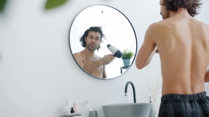 穿着睡衣的男人在浴室里用吹风机看着镜子吹干湿的长发