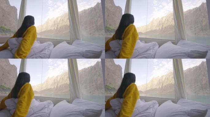 早晨醒来并透过湖边的窗户看时，穿着黄色衣服并包裹在温暖的毯子中的年轻女子的后视图。种族多样化的旅行者