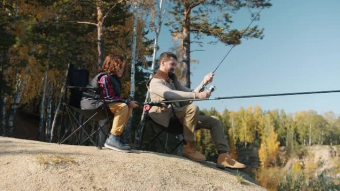 年轻的垂钓者与小儿子坐在椅子上抓着鱼竿，在外面享受秋天的日子