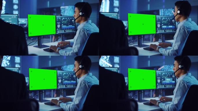 自信的男性数据科学家在大控制和监控室戴着耳机的绿屏模板的个人电脑上工作。年轻的工程师和同事在呼叫中心