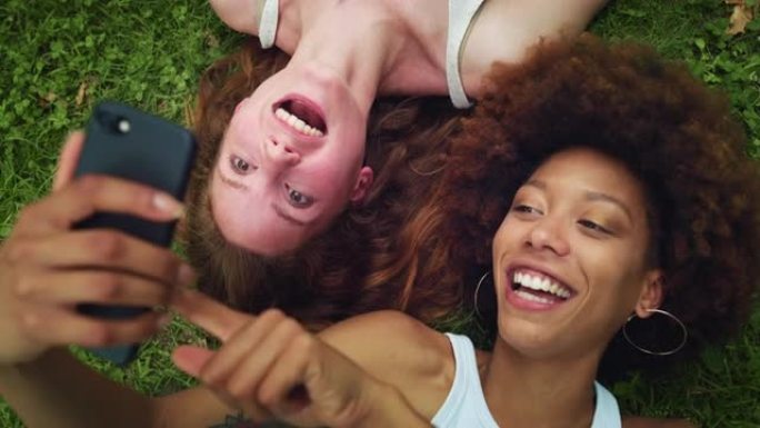 两名多民族女性躺在公园草坪上，一边用智能手机一边笑。不同的年轻女性最好的朋友在外面享受阳光明媚的温暖
