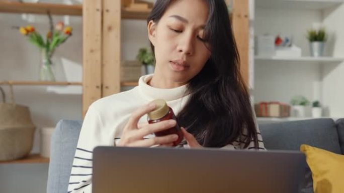生病的年轻亚洲妇女拿着药坐在沙发上视频通话用笔记本电脑在家咨询医生。