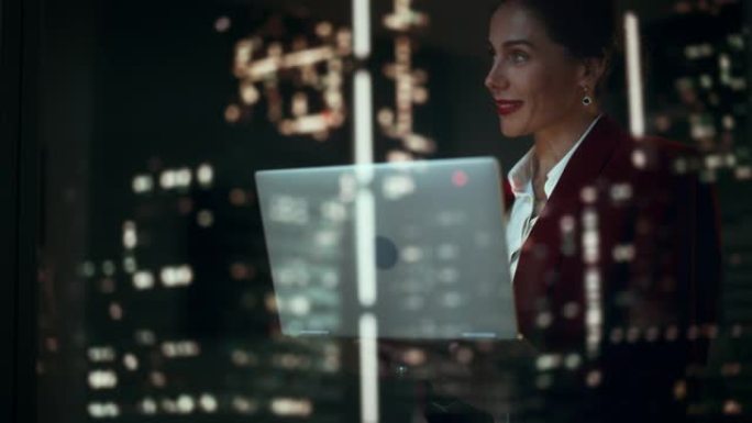 成功的女商人穿着时髦的西装，在笔记本电脑上看着夜城的奇迹。微笑的女首席执行官在计算机上工作到很晚，为