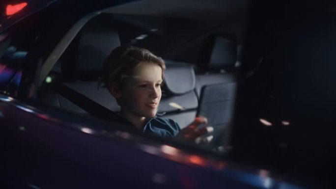 英俊的小男孩坐在汽车后座上，晚上通勤回家。乘客在平板电脑上玩视频游戏，而在城市街道的出租车上，霓虹灯
