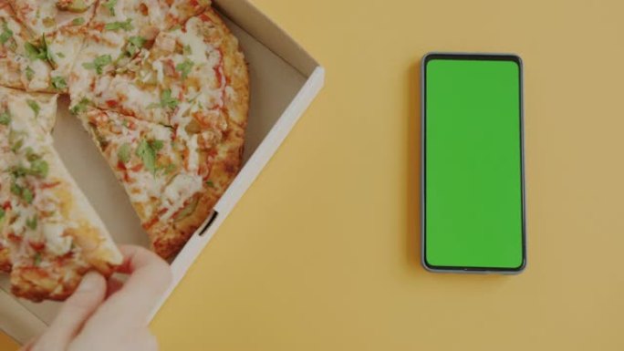 用绿屏色度键智能手机在黄色背景上拍摄美味披萨的手特写