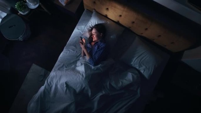 顶景卧室公寓: 美丽的年轻女子晚上在床上使用智能手机。微笑女孩浏览社交媒体，浪漫聊天，约会应用程序，
