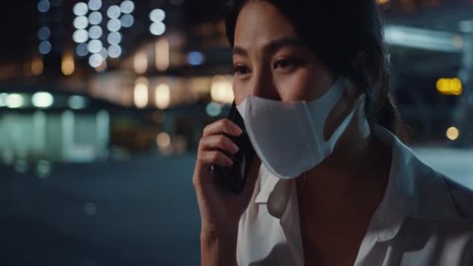 严重不满的年轻亚洲女商人戴着医用口罩通过电话交谈，独自一人在城市夜晚户外行走。