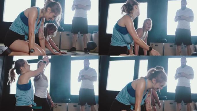 两名年轻快乐的白人妇女在锻炼后极度疲倦，在健身房地板上呼吸沉重，互相击掌。
