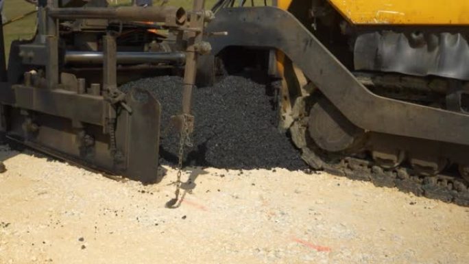 特写: 重型摊铺机机械将新鲜的黑色沥青滴在碎石上