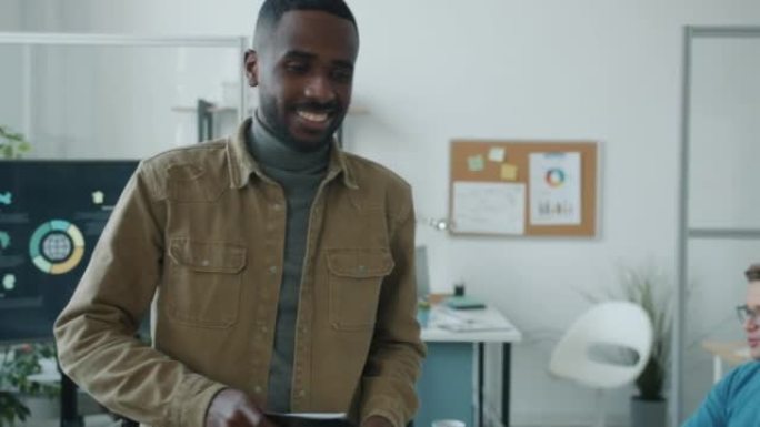自信的非洲裔美国商人走进办公室与同事交谈，然后使用笔记本电脑