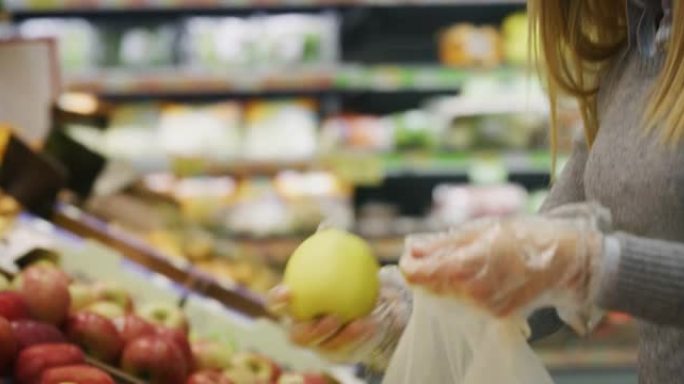 戴着医用口罩和手套保护自己免受疾病侵害的年轻女子的真实特写镜头正在超市购物时选择苹果。