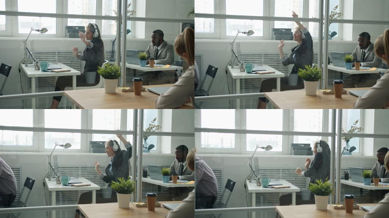 快乐的女上班族戴着耳机跳舞，坐在共享工作场所的办公桌前玩得开心