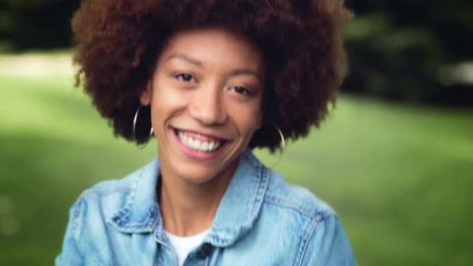 特写一位年轻黑人妇女看着相机在绿色公园跳舞的肖像。快乐的卷发女性享受新鲜的空气和自然，与世界分享她的