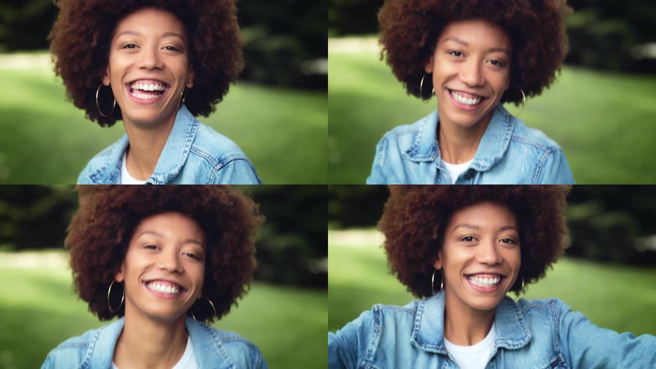 特写一位年轻黑人妇女看着相机在绿色公园跳舞的肖像。快乐的卷发女性享受新鲜的空气和自然，与世界分享她的