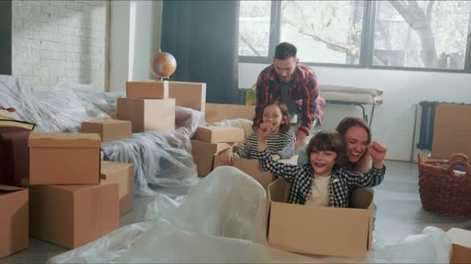 搬进新家。年轻快乐的父母在新宽敞的房子地板上把可爱的孩子推到纸箱里很开心。